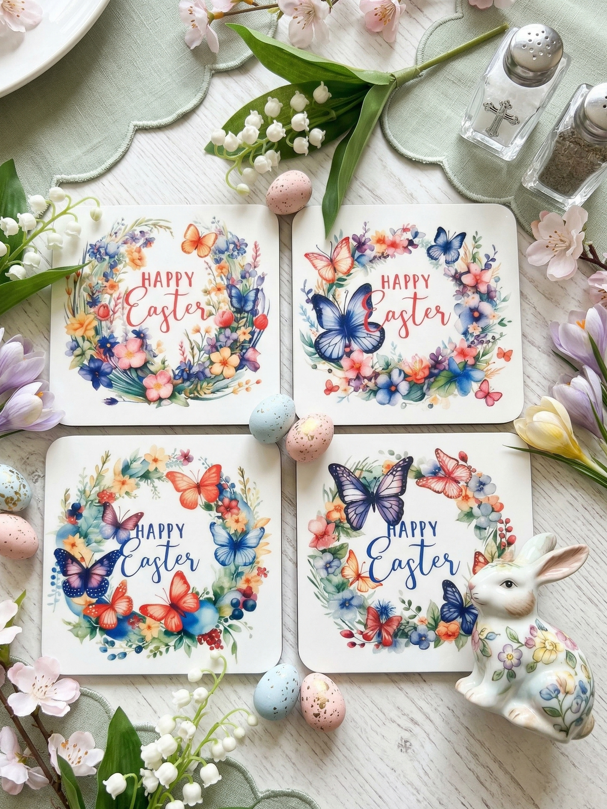 Four square mats with floral wreaths on white wood with colourful eggs, a ceramic rabbit, and lily of the valley.