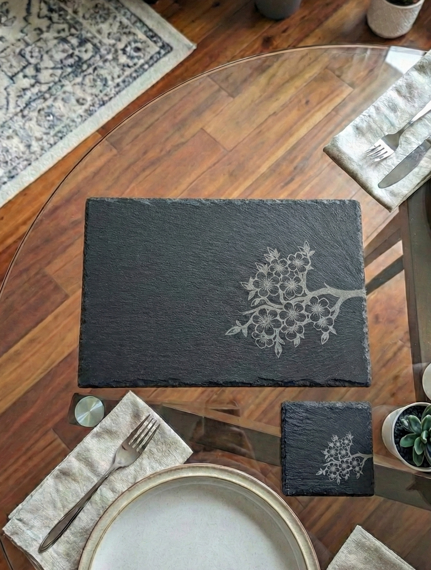 Black slate placemat with a grey branch engraving, shown with a white plate and silver fork on a glass table.