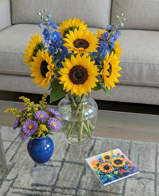 Sunflowers in a clear vase and purple flowers in a solid blue vase on a glass table with one floral coaster.