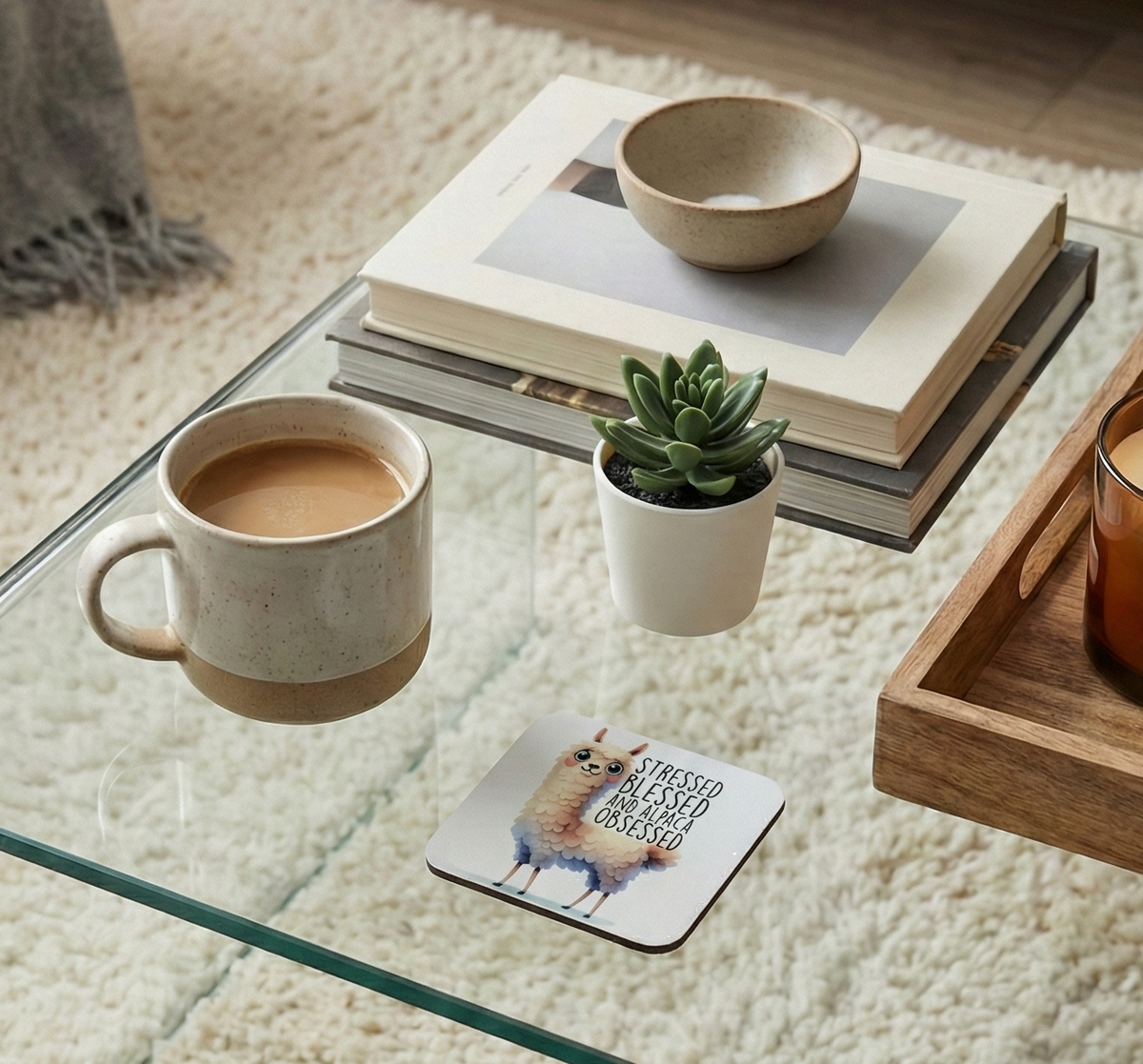 Colourful alpaca coaster on a glass table with a ceramic mug and a wooden tray containing a candle nearby.