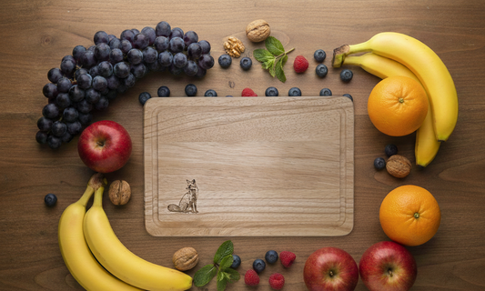 Flat lay of light wood board, engraved fox detail, on dark wood background. Framed by grapes, bananas, red apples, oranges, and berries.