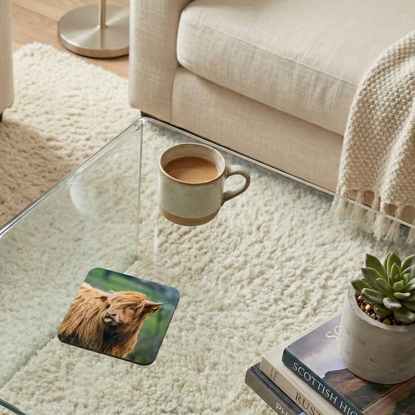 Square coaster with a ginger Highland cow pulling a face on a glass table. Cream mug and beige textured sofa.