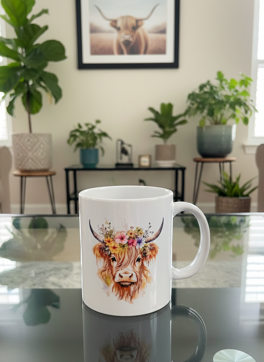 White ceramic mug with a watercolour Highland cow and floral crown sits on a dark, reflective table. Potted green plants and framed artwork in background.