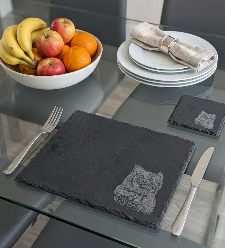 Dark grey textured slate placemat with an owl etching. Silver cutlery, white plates and colourful fruit on a glass table.