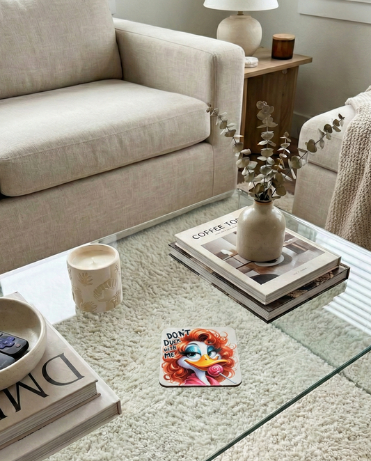 Close-up of a glass coffee table over a cream shag rug. Features a vase with dried branches, books, and a novelty red-haired duck coaster.