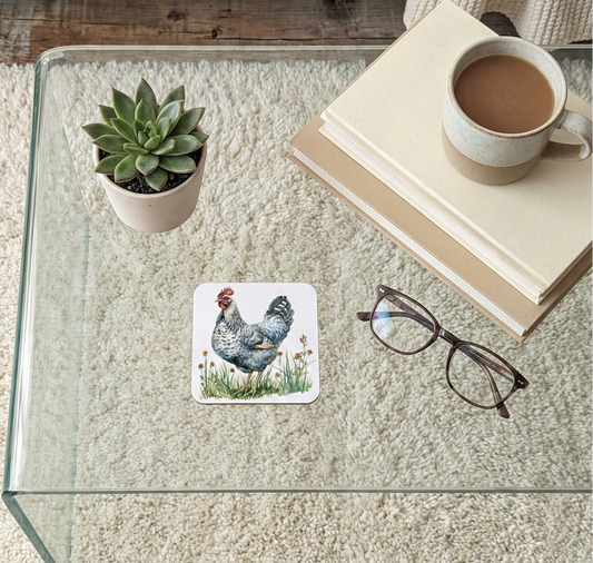 Close-up of a square coaster showing a speckled Grey chicken standing in a meadow illustration on a glass table.