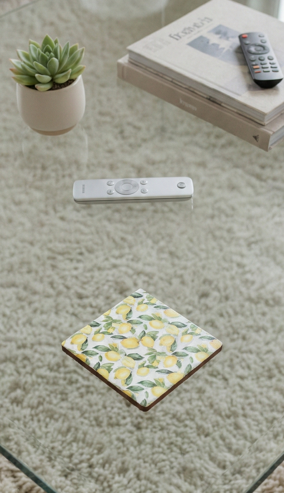 Square coaster with a yellow and green lemon pattern on a glass table above a shaggy cream-coloured rug.