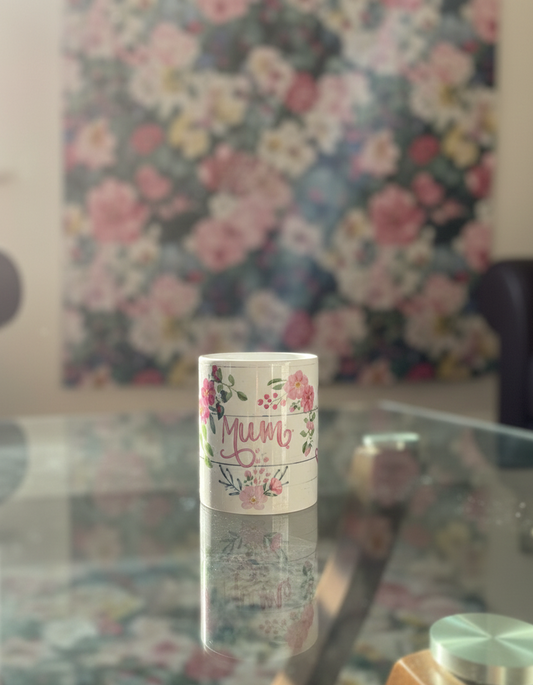 A "Mum" floral mug sitting on a glass coffee table in a sunlit living room. The mug's reflection is visible on the glass surface, and the background features a soft-focus floral wall hanging and warm, natural light.
