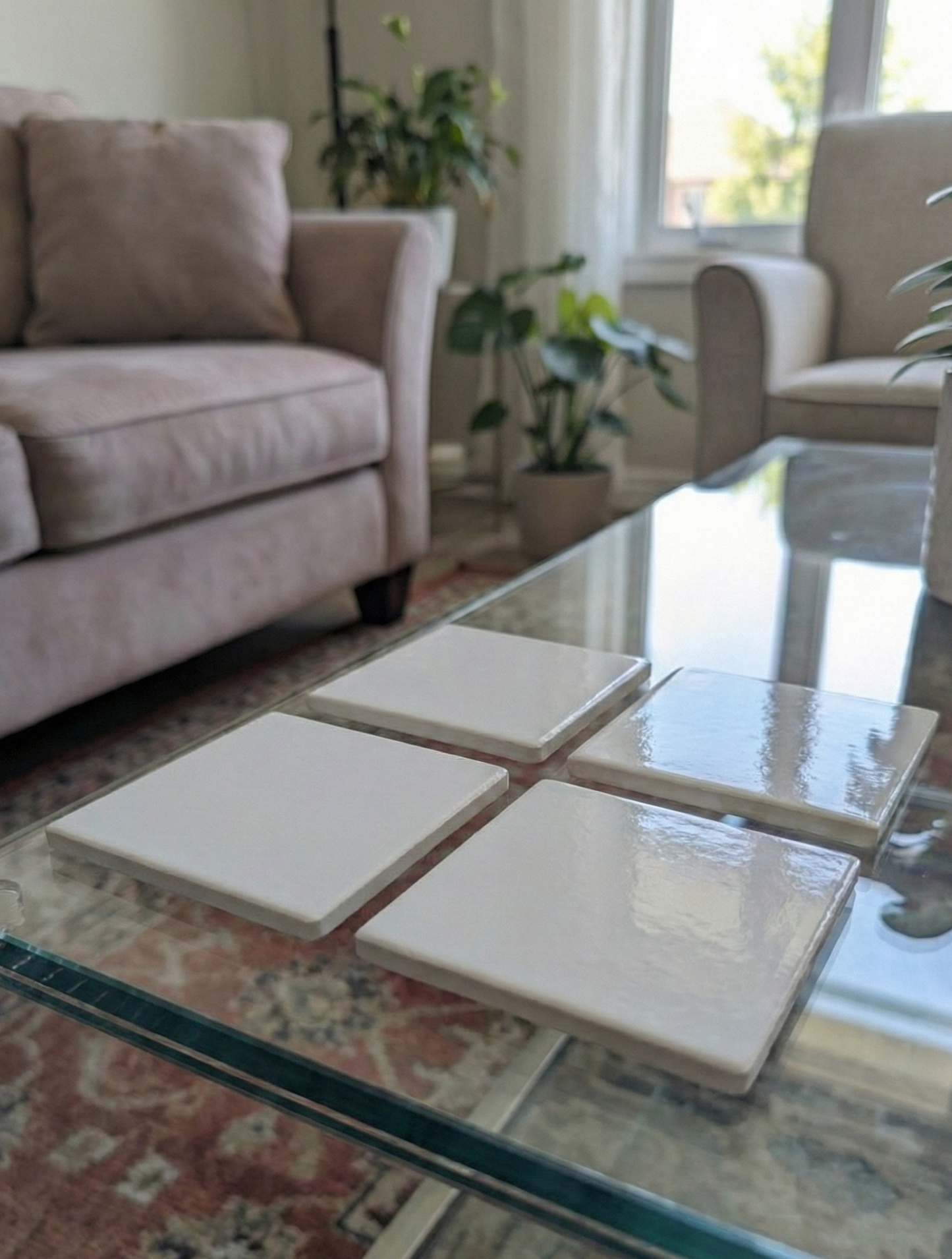 Four highly reflective glossy white square coasters arranged in a grid on a clear glass coffee table with a patterned rug.