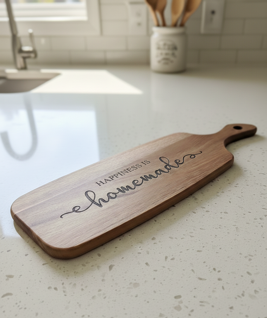 Long, brown wooden paddle board, etched with "Happiness is homemade" script, lying flat on a bright speckled white countertop.