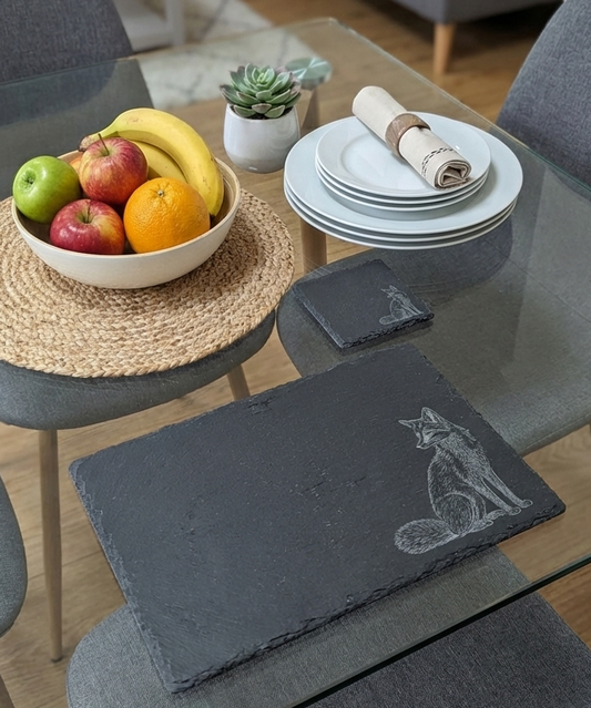 Rectangular dark grey slate placemat with an etched fox. Round woven mat, colourful fruit bowl and white plates nearby.