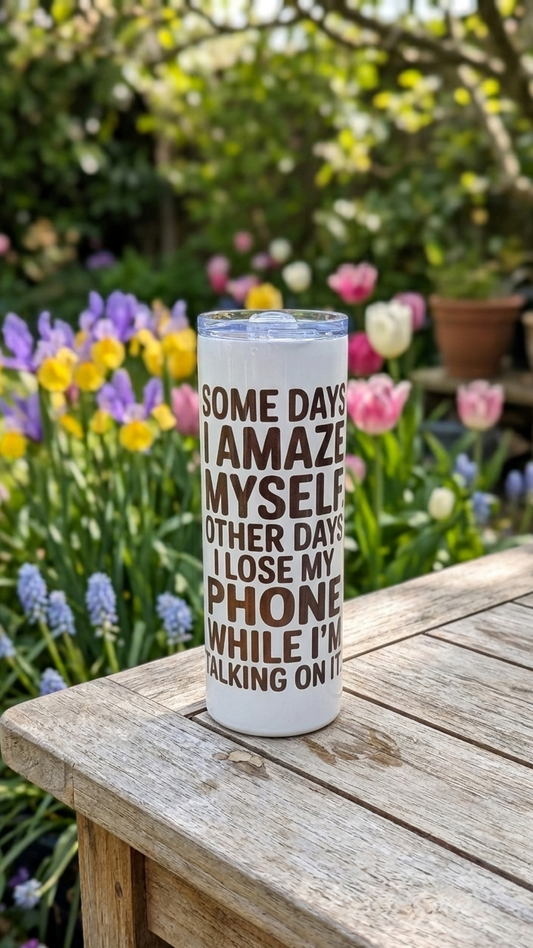 Tall white tumbler with brown text on a grainy wooden table. Background shows colourful purple, yellow and pink flowers.