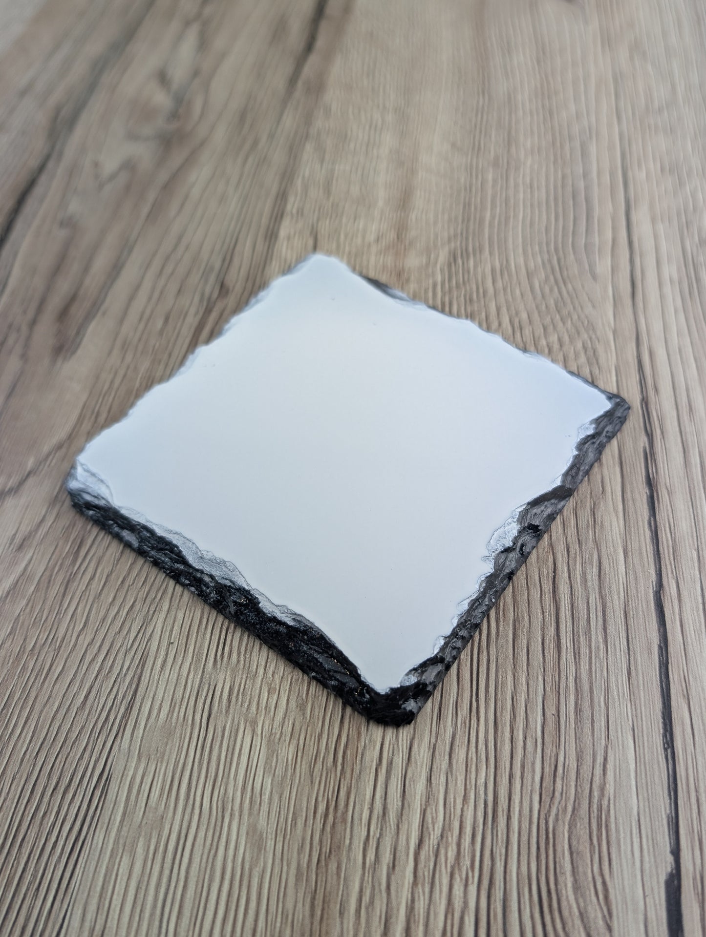 Square slate coaster with a smooth white top and jagged dark grey edges, set on a light wood-grain background.