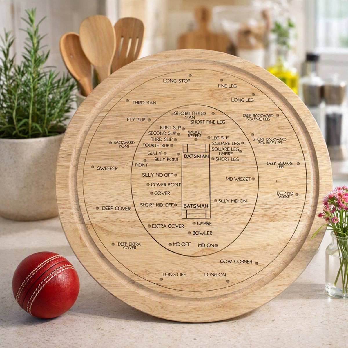 Engraved wooden cricket field placement board on a kitchen worktop with utensils, herbs, flowers and a red cricket ball.
