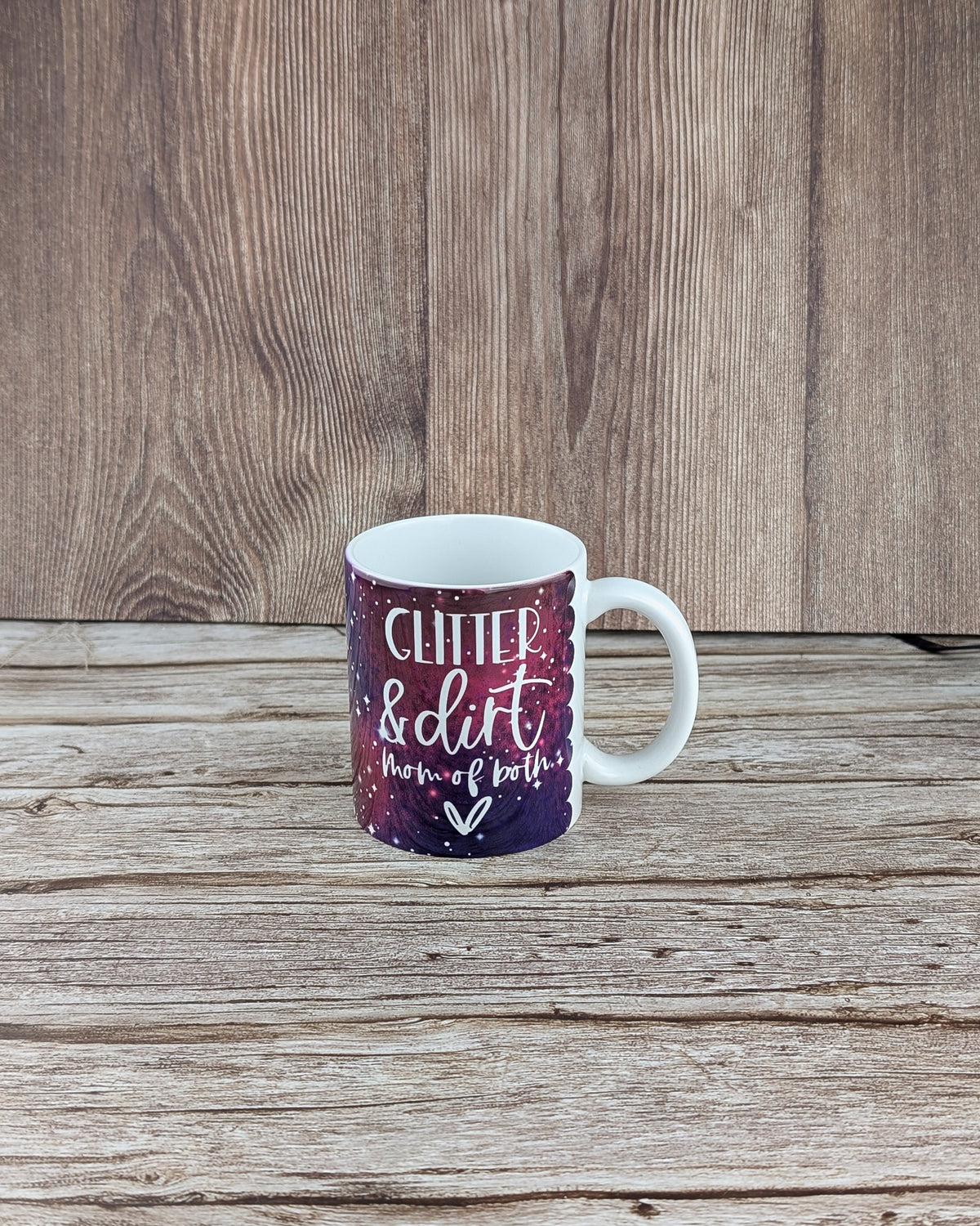 White ceramic mug features a purple, magenta, and white galaxy print. Resting on grained wooden planks with a dark wood wall background.