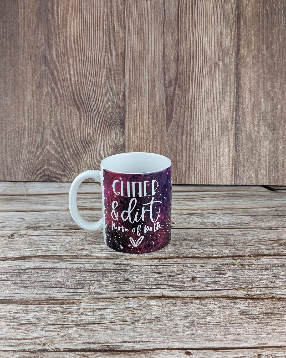White ceramic mug features a purple, magenta, and white galaxy print. Resting on grained wooden planks with a dark wood wall background.