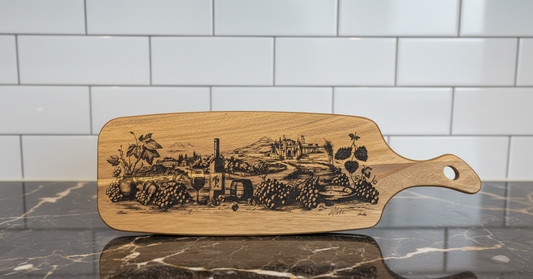 Front view of a rectangular wooden charcuterie board with a handle, featuring a detailed black ink-style engraving of a vineyard landscape, grapes, and wine bottles, resting on a dark marble countertop with a white subway tile backsplash.