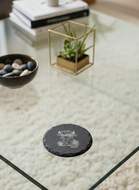 Circular dark grey slate coaster with a textured edge, etched with a sitting Highland cow, on a glass table.