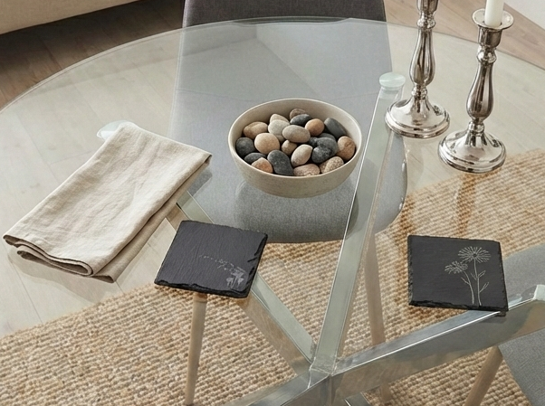 Two square charcoal slate coasters with floral engravings on a glass table beside a bowl of natural grey pebbles.