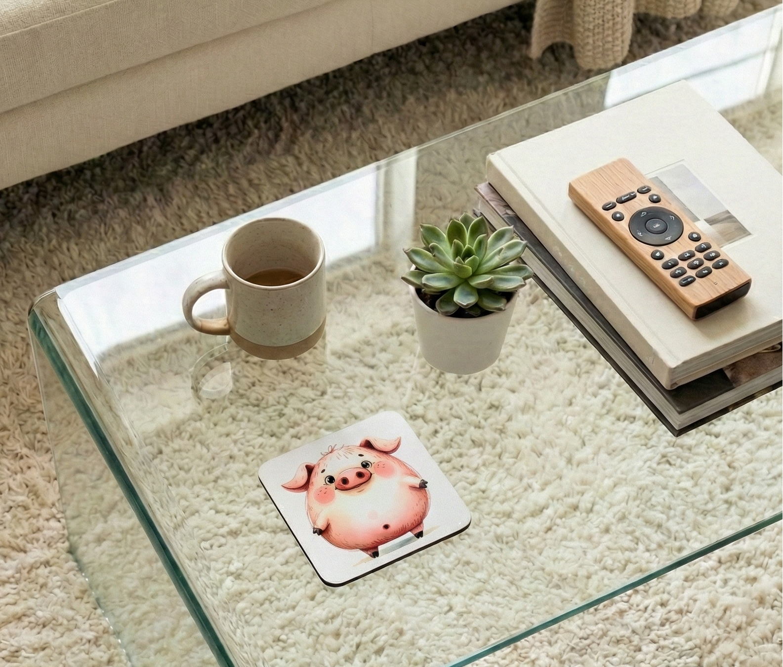 Square white coaster with a round pink pig illustration on a glass table next to a cream mug and green succulent.