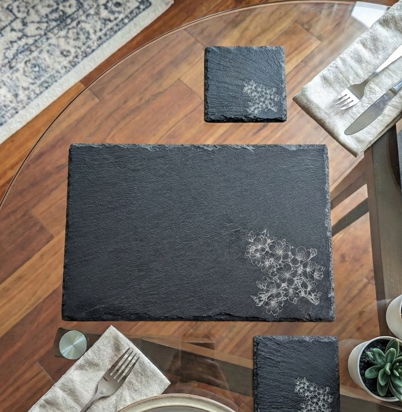 Top-down view of a black slate placemat with a grey floral cluster engraving, silver cutlery, and beige linen napkins.