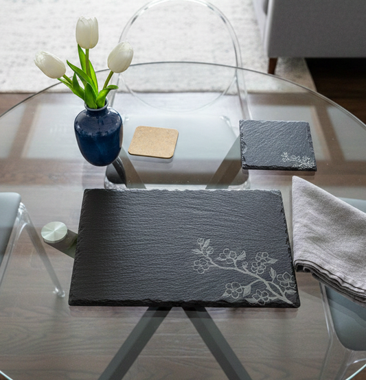 Black slate placemat with a grey floral engraving on a glass table with white tulips in a blue vase.