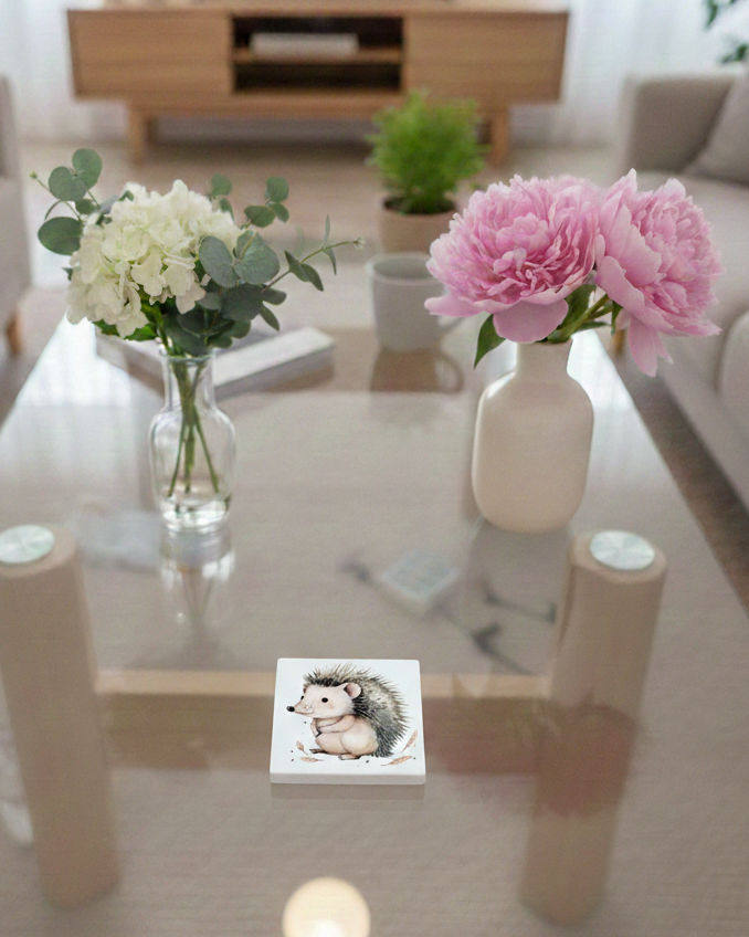 White square coaster on glass table showing a hedgehog side-profile illustration with cream and earthy brown colours.