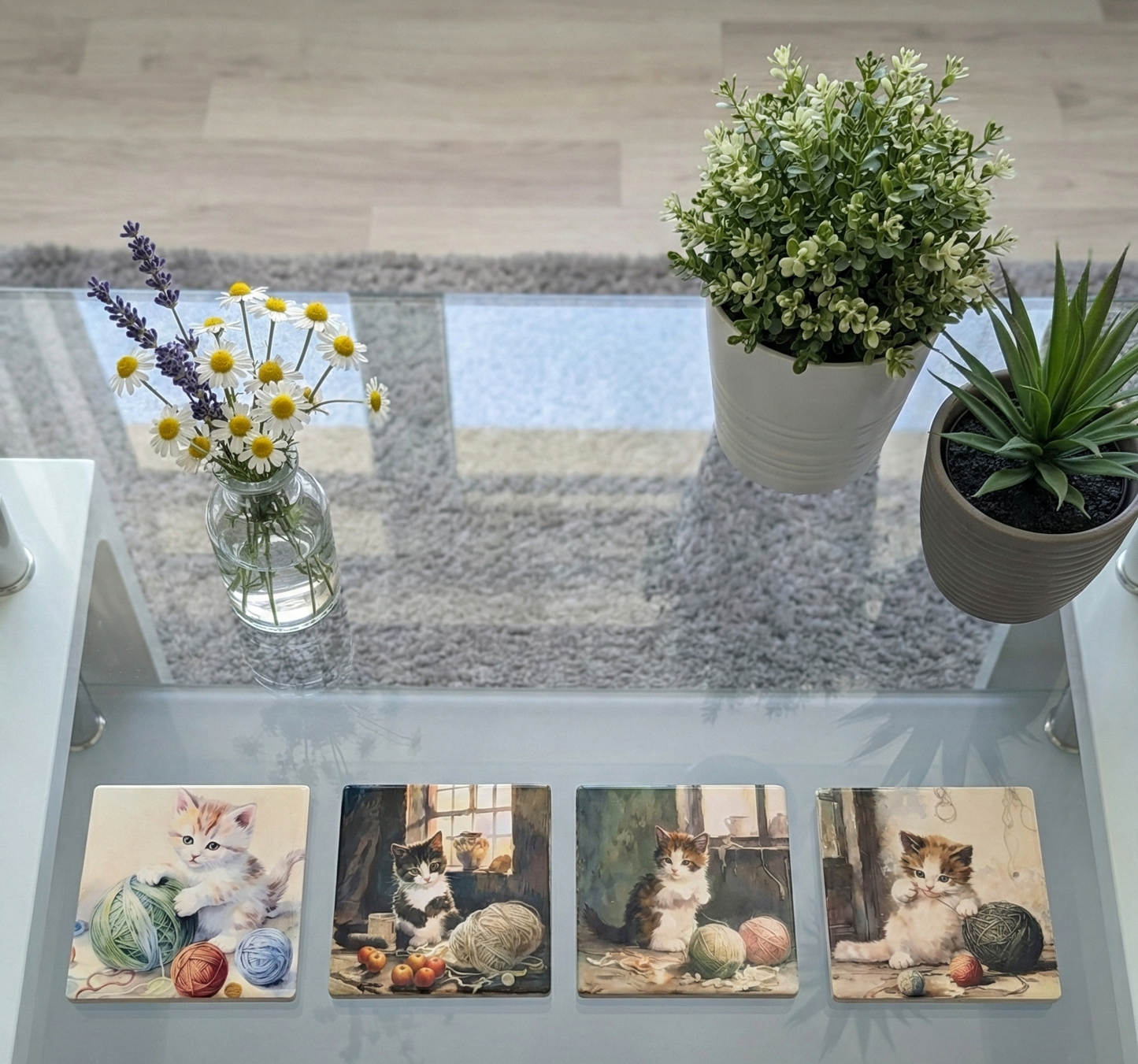 Four kitten coasters on a glass table with white daisies, lavender, and green plants in white and grey pots.