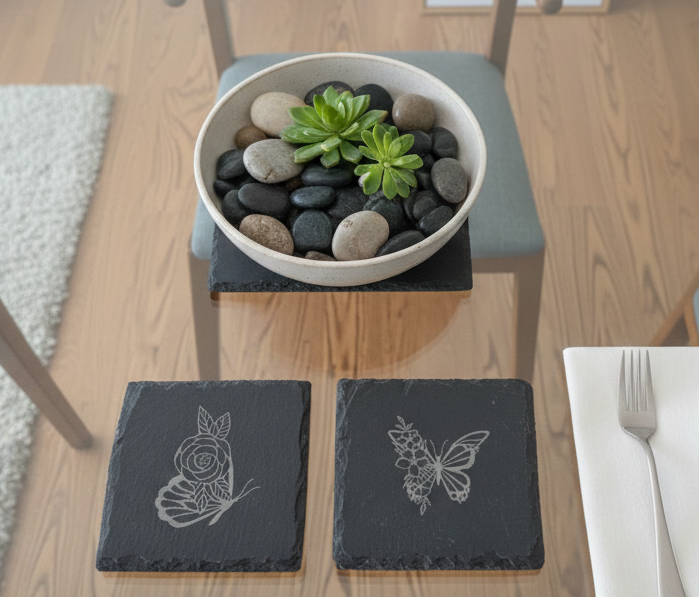 Overhead shot of two square slate coasters with butterfly and rose engravings on a glass coffee table.