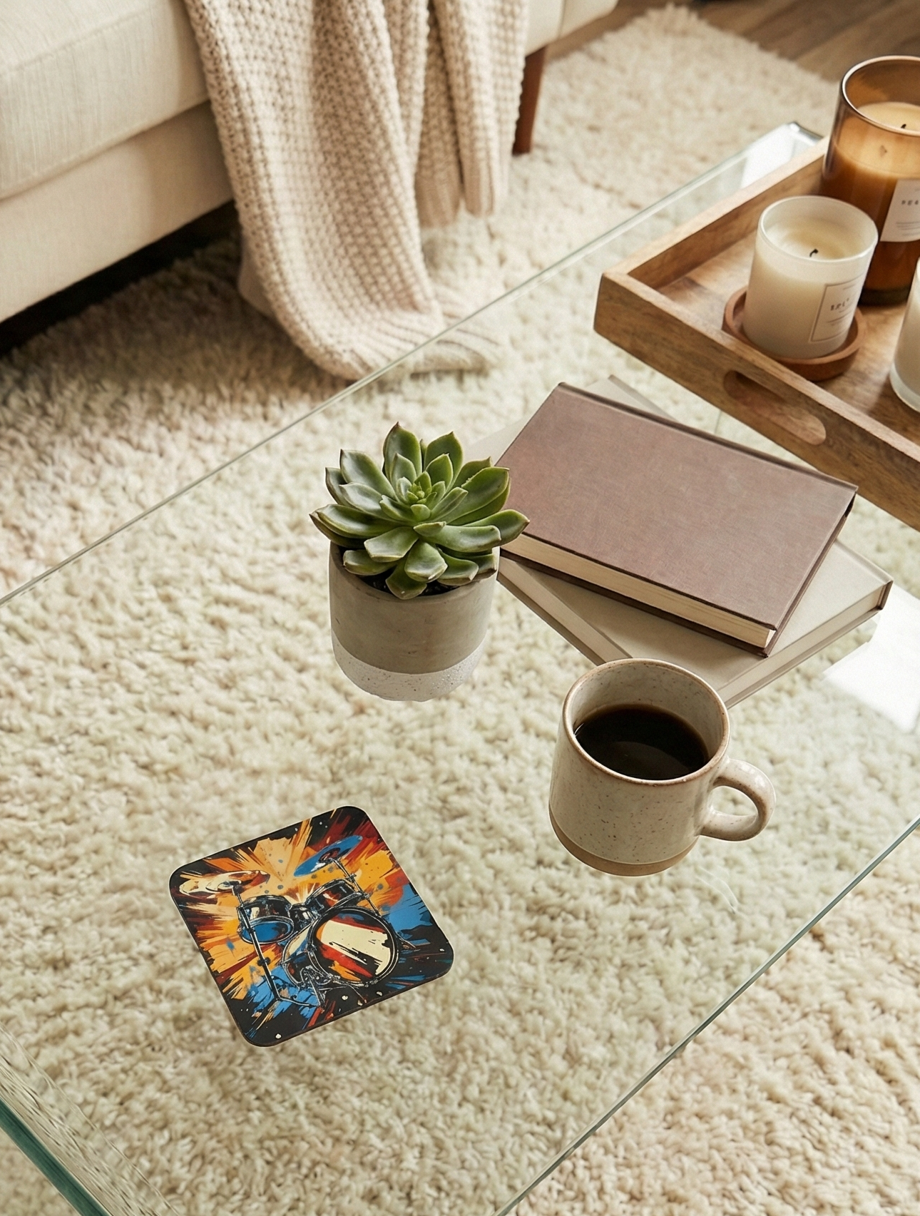 Overhead view of a glass table on a high-pile beige rug. Features a succulent, books, coffee, and a colourful, abstract drum coaster.