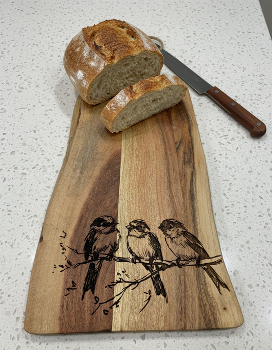 Smooth wooden serving board with warm brown grain and etched birds, holding a crusty white sourdough loaf and knife.