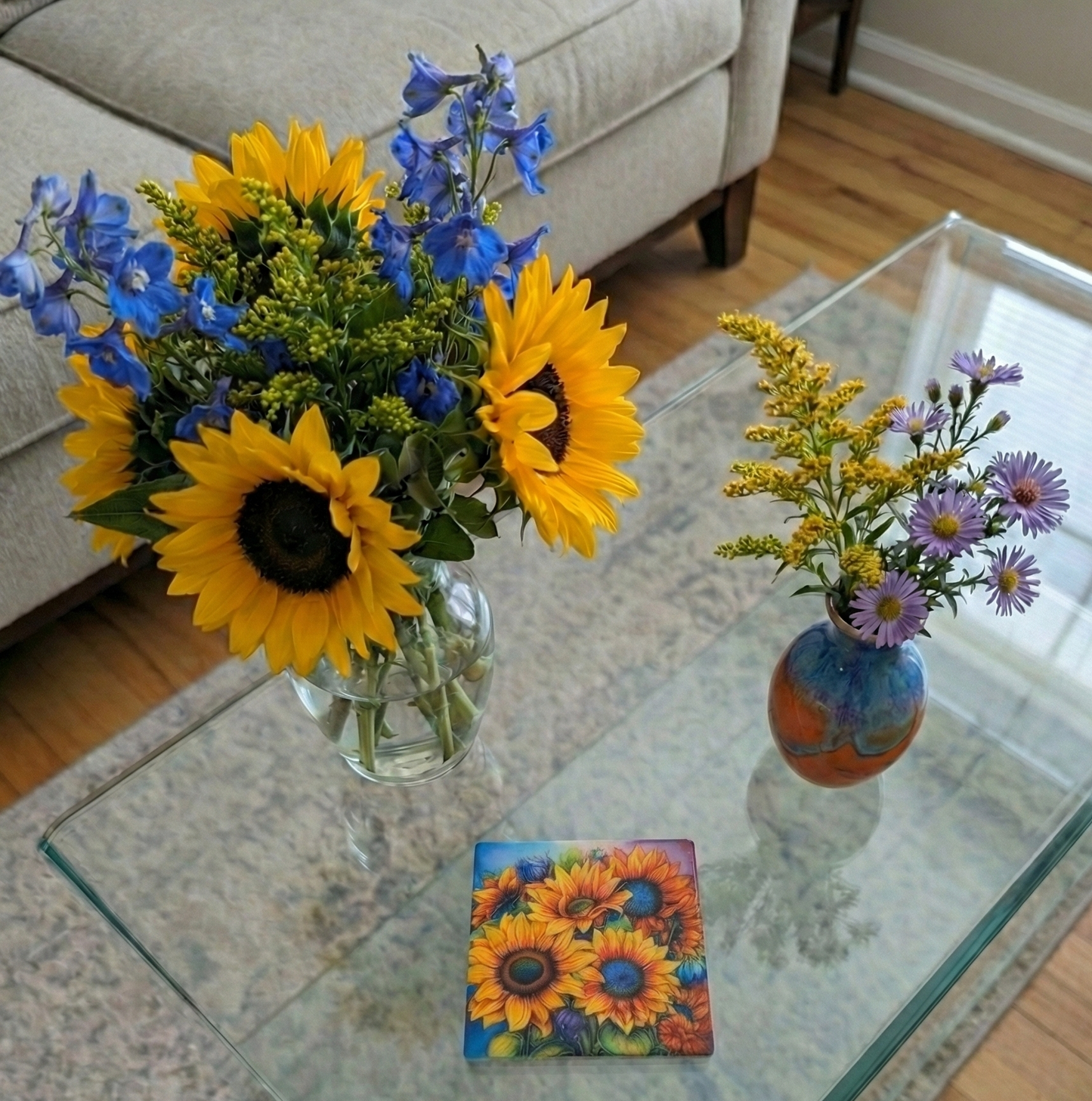 High-angle view of sunflowers and blue delphiniums next to an orange and blue vase and a square coaster.
