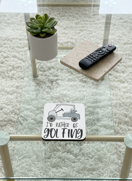 Square white coaster with a grey golf cart illustration and text, on a clear glass table over a white shaggy rug.
