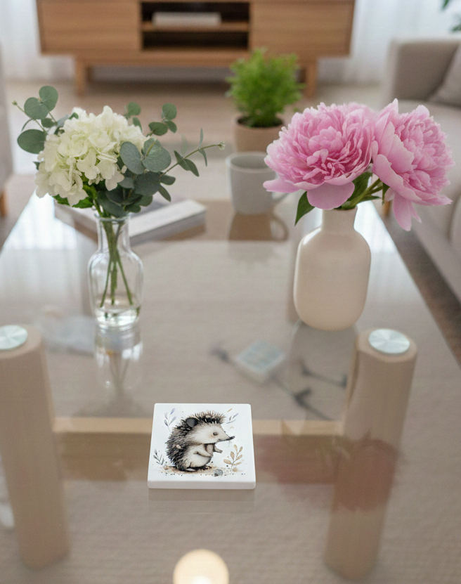 White square coaster on a glass table with a hedgehog profile illustration amongst delicate grey and cream leaves.