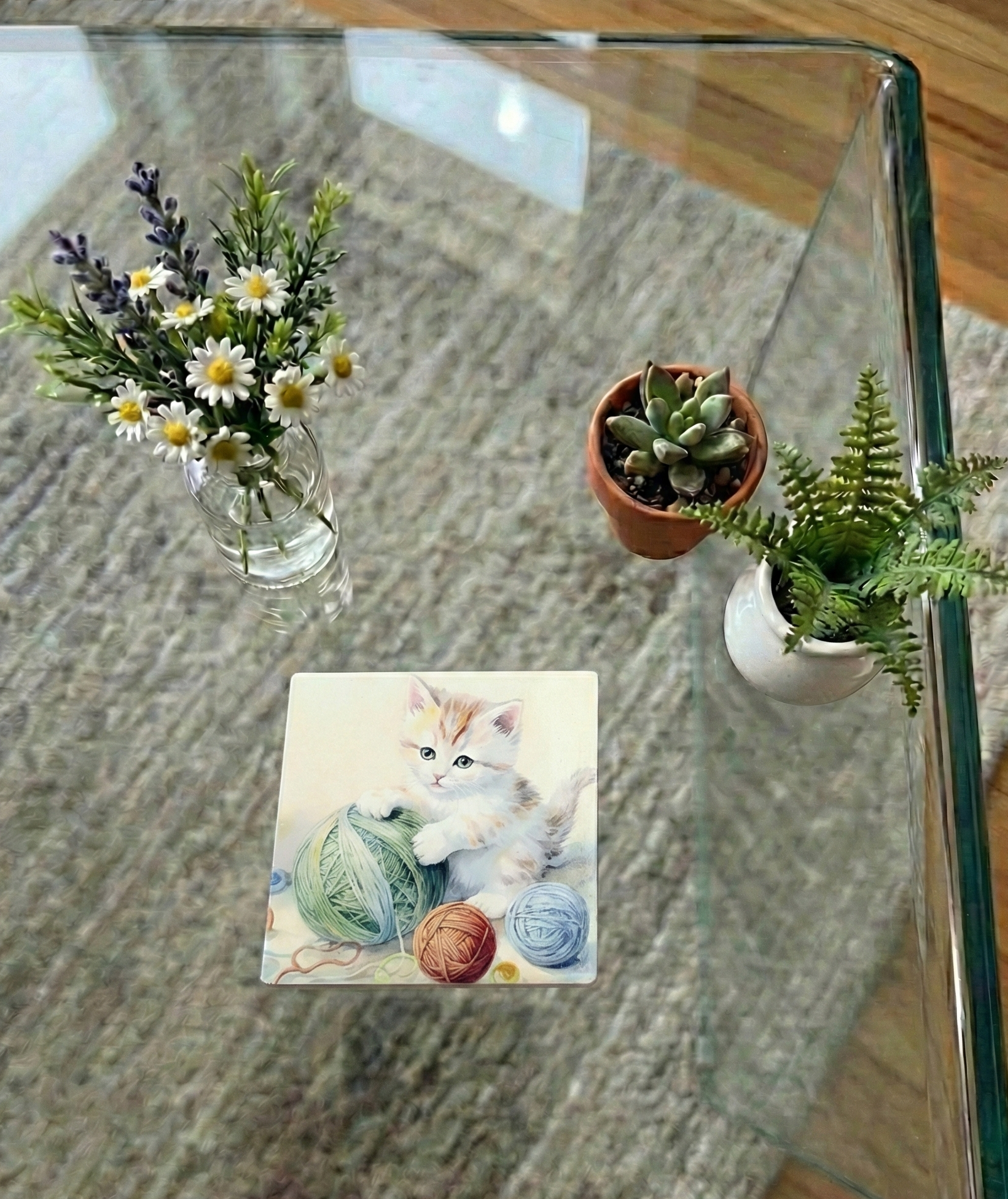 Ginger and white kitten coaster with a large green ball of wool on a glass table beside daisies and lavender.