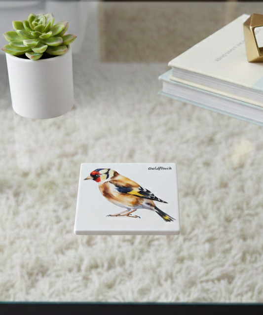 Glossy white square coaster with a colourful Goldfinch illustration, resting on a glass table over a cream, shag rug.