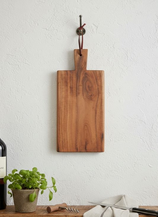 Rich brown wooden cutting board hangs on a textured white wall above a wooden shelf holding a wine bottle and green basil.