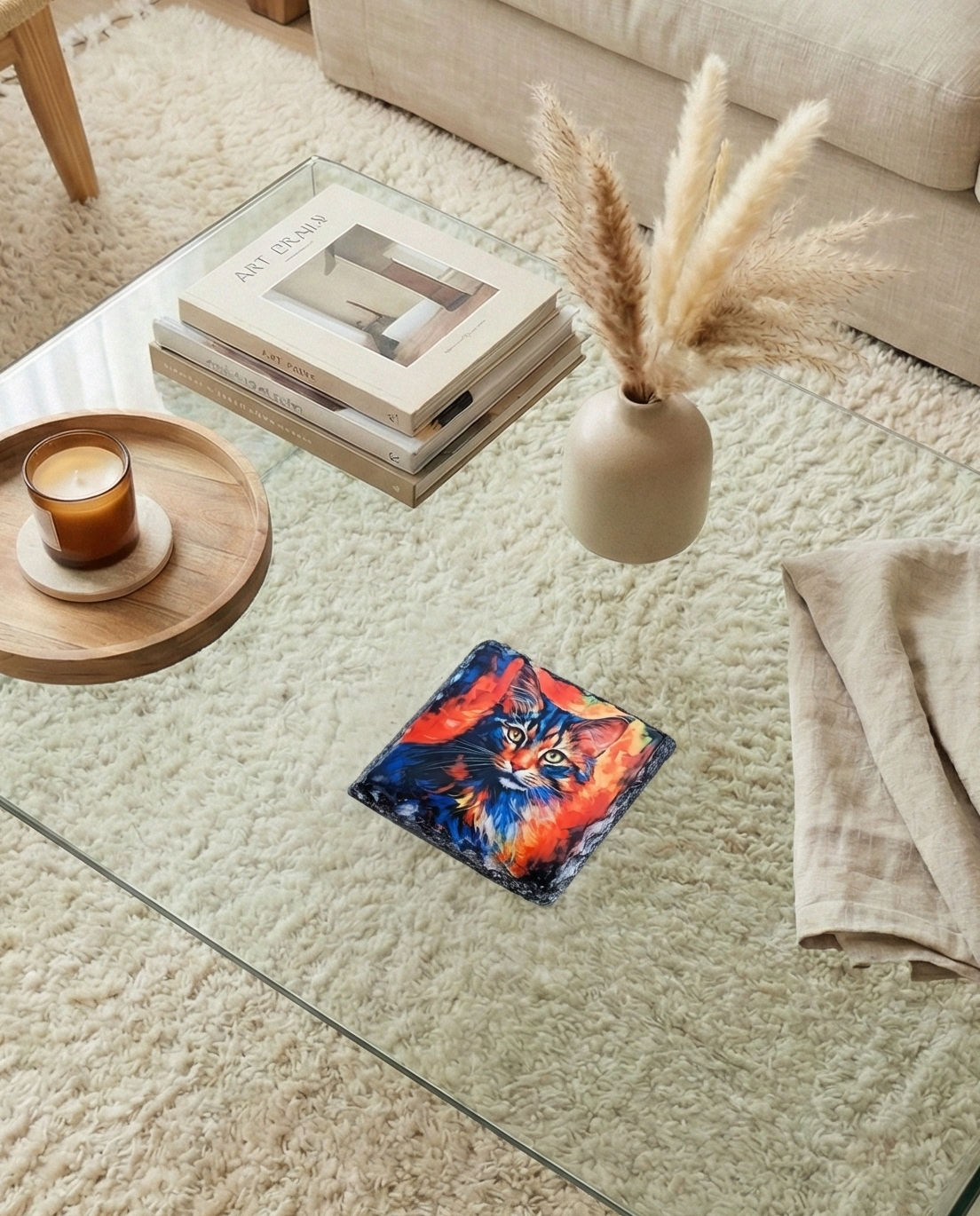 A square slate coaster featuring a dark, richly colourful portrait of a fluffy cat with striking blue eyes, on a glass surface.