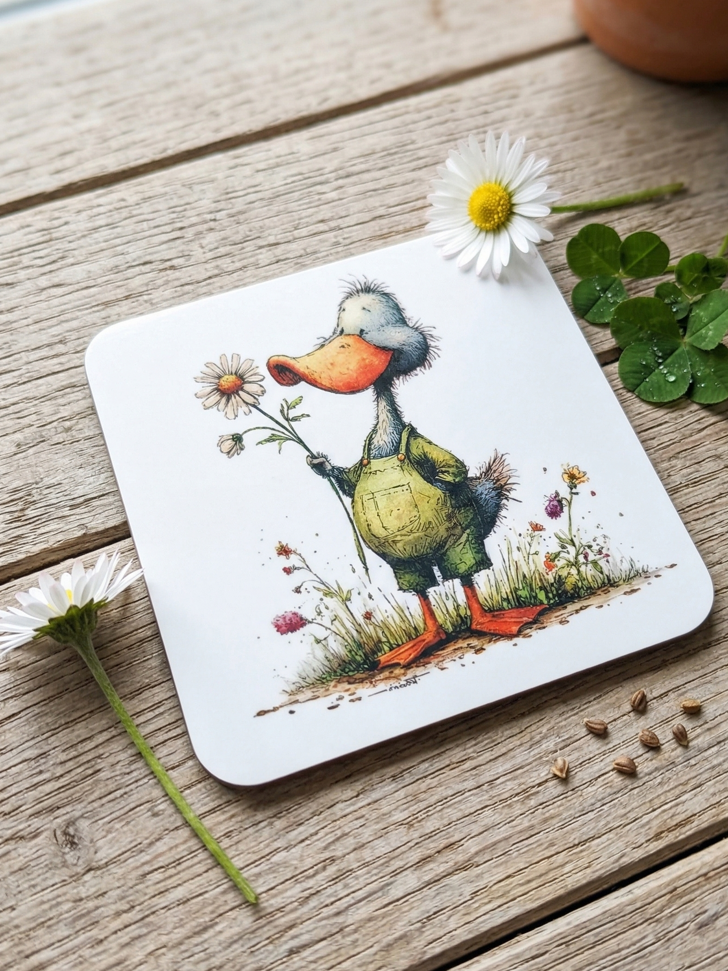 Close-up of a duck coaster on a wooden surface, beside a fresh white daisy, green clover, and brown seeds.