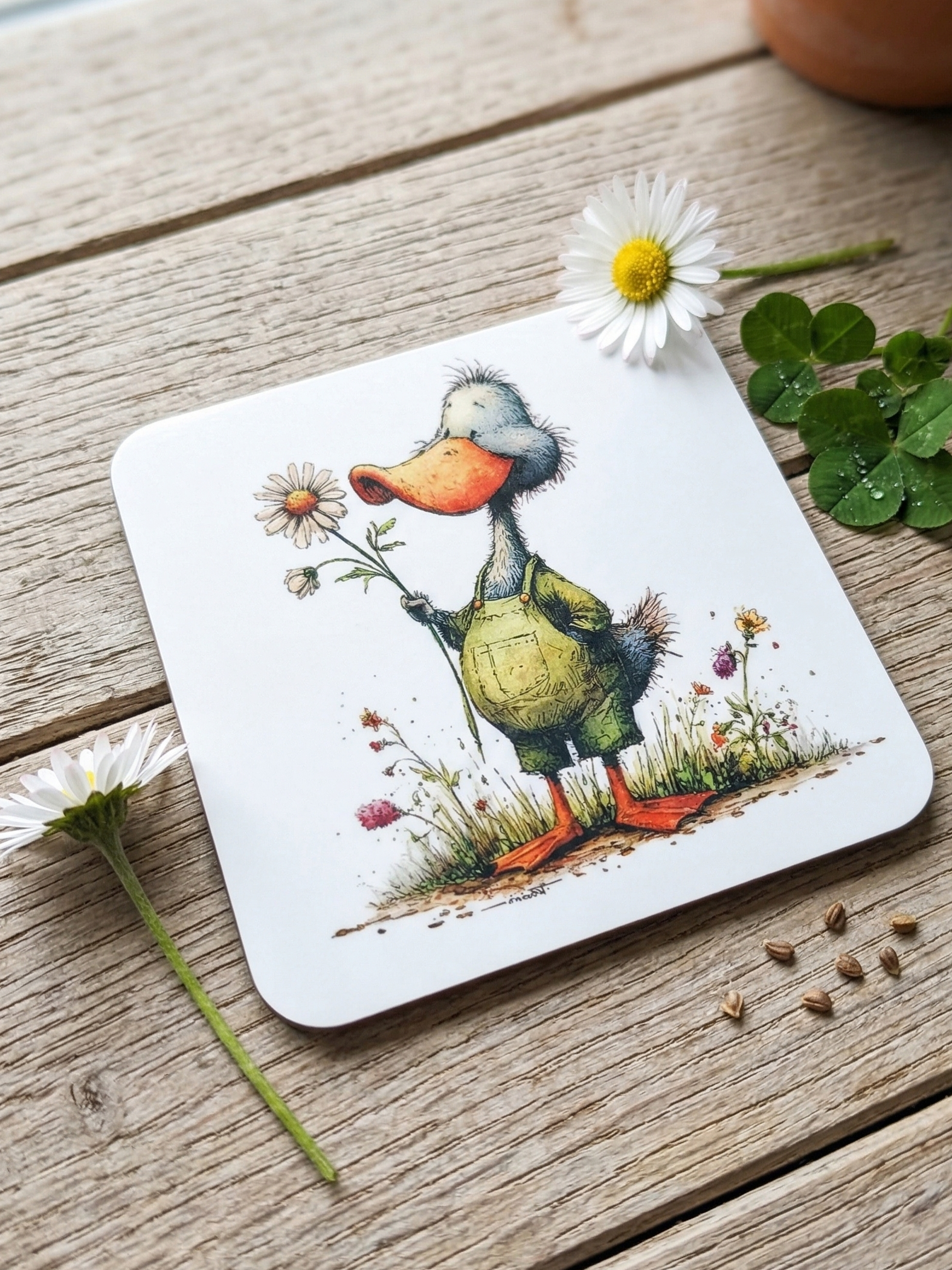 Close-up of a duck coaster on a wooden surface, beside a fresh white daisy, green clover, and brown seeds.