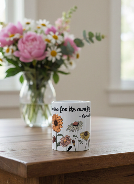 White ceramic mug detailed with colourful retro flowers and quote, on a thick wooden table; blurred pink peony bouquet in background.