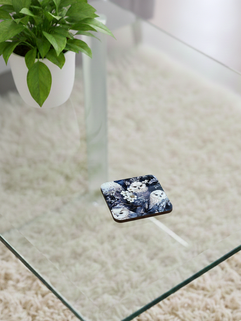 A square coaster featuring white owls and blossoms on a blue-grey background, resting on a transparent glass surface.