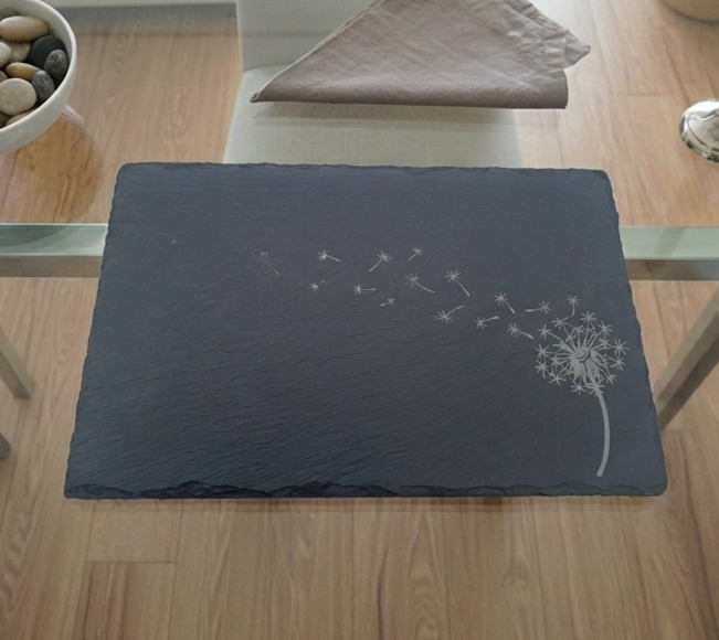 Charcoal grey slate placemat featuring a detailed white dandelion engraving with seeds floating across the surface.