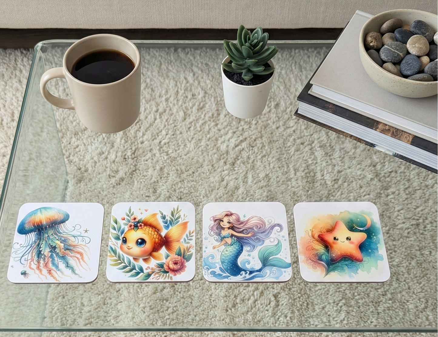 Four coasters with sea creature illustrations on a glass table beside a beige mug, a green succulent, and grey stones.