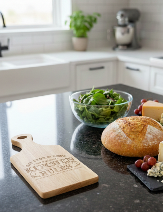 Light engraved wooden cutting board and rustic bread on a dark, glossy, speckled granite countertop. Grapes and cheese nearby.