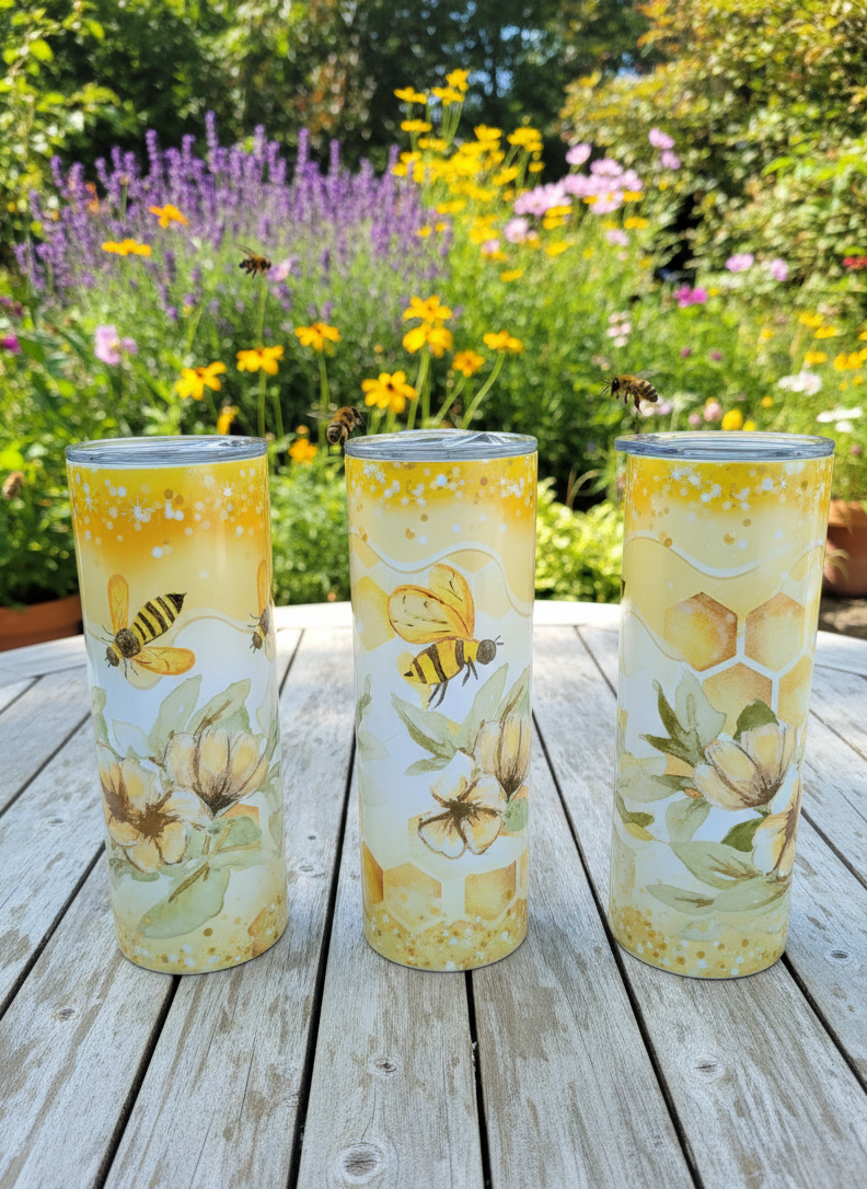 Three tall tumblers with honeybee and floral designs on a wooden garden table, surrounded by colourful summer flowers.