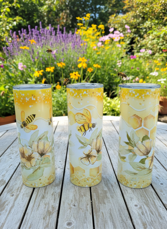 Three tall tumblers with honeybee and floral designs on a wooden garden table, surrounded by colourful summer flowers.