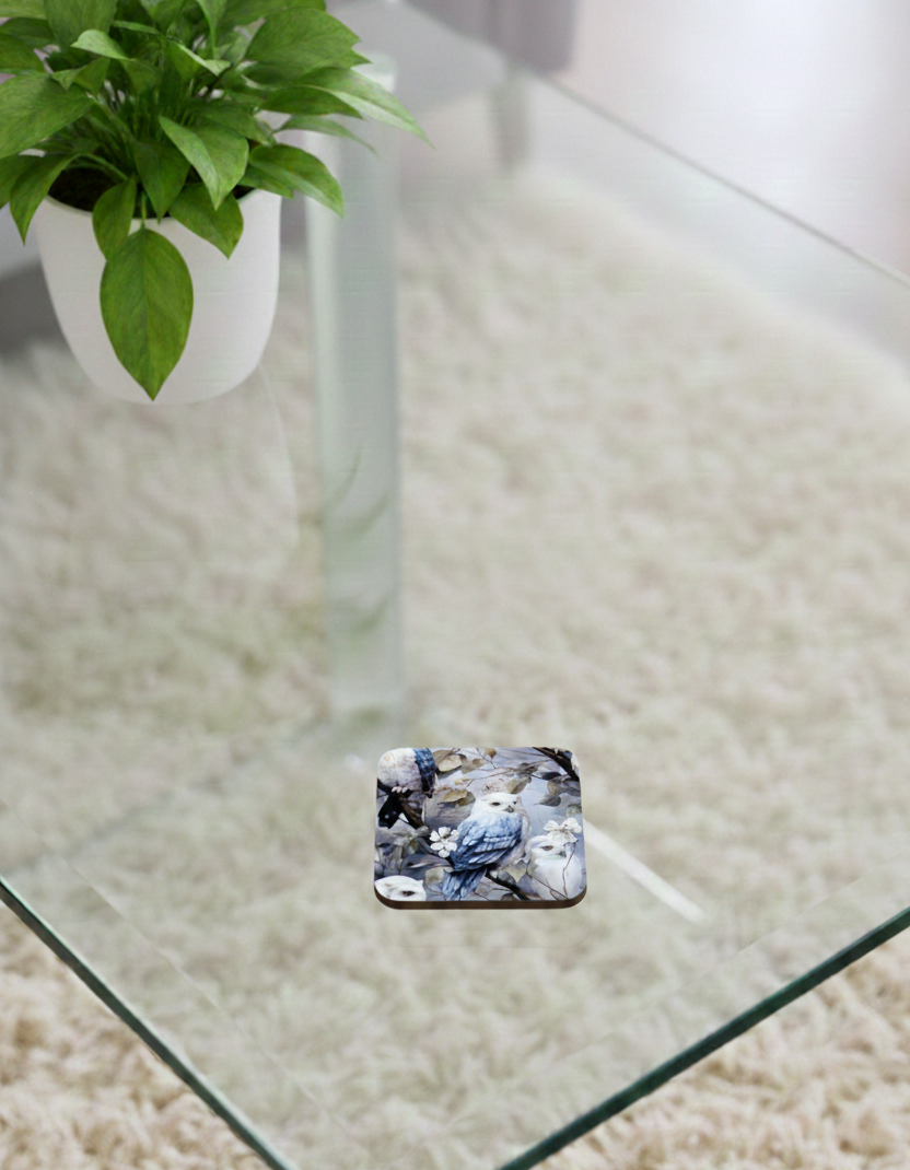 Square coaster with a blue-grey owl perched on branches, shown on a glass coffee table over a neutral shaggy rug.