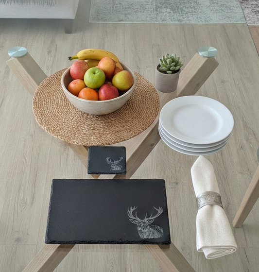 Dark grey slate placemat with a white etched stag. Round woven mat with fruit bowl and white plates on a glass table.