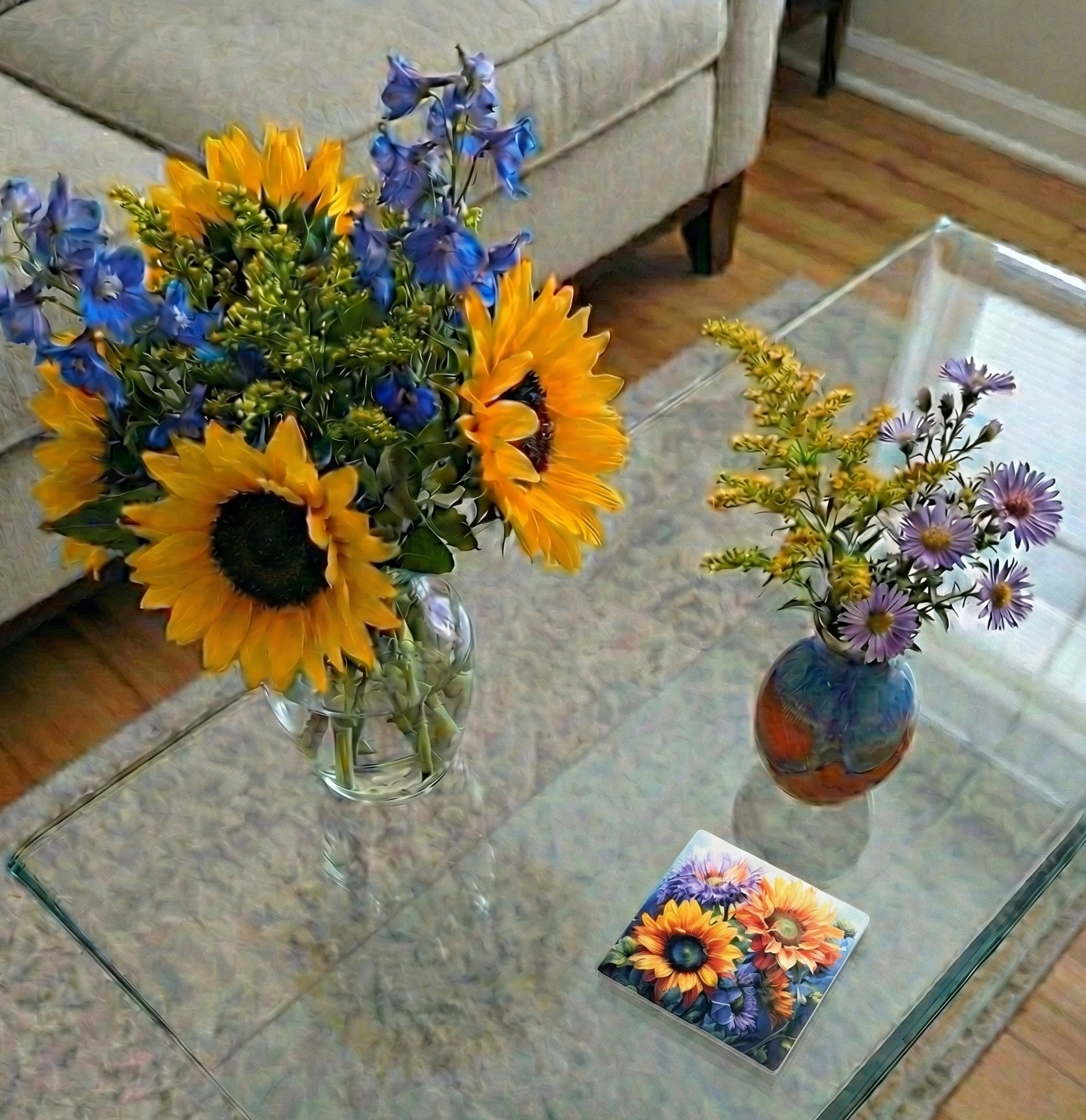 A stylised, painterly shot of sunflowers and delphiniums on a glass table with a single floral coaster.